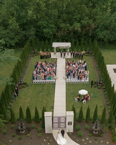 Aerial view of an outdoor wedding ceremony in Ohio. Rows of seated guests face a flower-adorned gazebo, with trees surrounding the elegant garden setting.