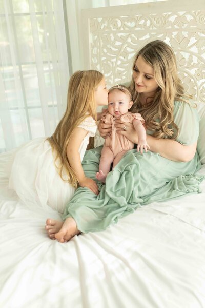 Mother sits on a white bed with her baby while big sister leans in for a sweet kiss on the baby’s head.