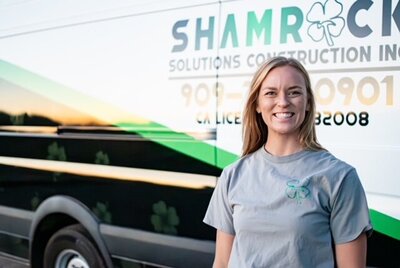 A blonde woman wearing a grey shirt smiling in front of a Shamrock Solutions Construction Inc van.