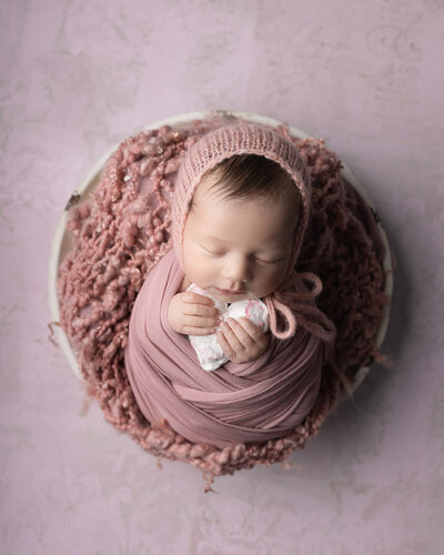 Sleeping newborn girl in light pink wrap and bonnet, with pink background, holds tiny white heart in her hands