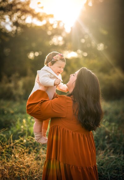 A mom in a orange dress is holding her baby up while the sun sets.
