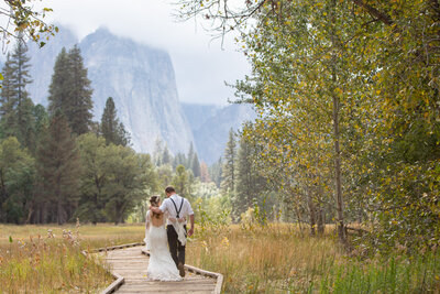 A newlywed couple walks along a wooden boardwalk in Yosemite Valley, with El Capitan towering in the misty background. Captured by Yosemite elopement photographers, this peaceful moment reflects the beauty of an intimate wedding surrounded by nature.