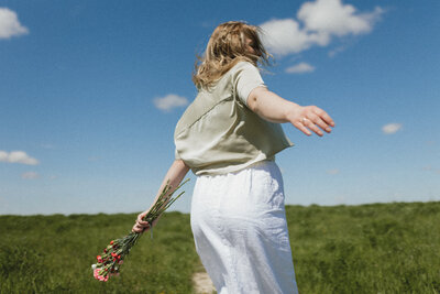 A young girl runs joyfully through a grassy field, holding a bouquet of flowers.