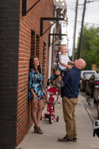 A father lifts his laughing toddler in a playful moment on a sidewalk. A smiling woman leans against a brick wall nearby. A red tricycle is behind them.