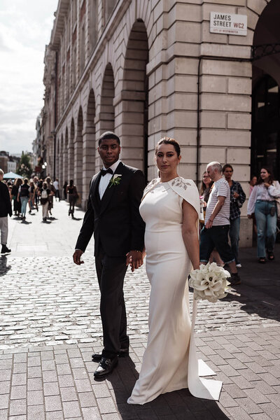 bride holding a bouquet of calla lilies in one hand and her groom’s hand walking through the streets of london to the nomad hotel for their intimate wedding
