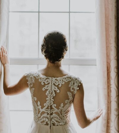 photo of a bride in her wedding dress looking out the window of her bridal suite as light comes pouring in.