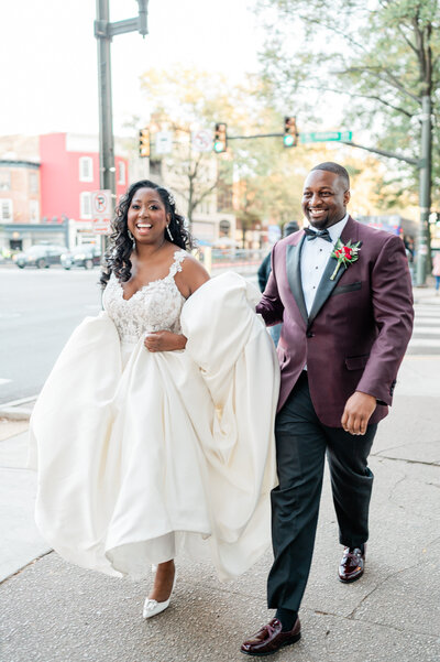 A groom helping a bride hold the bottom of as they walk down a sidewalk