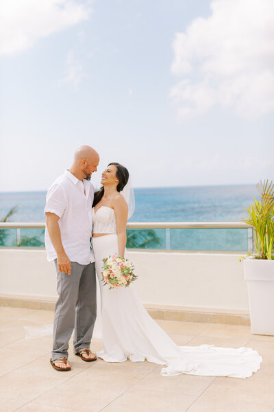 A wedding couple embraces on a tropical rooftop terrace with modern buildings and palm trees in the background.