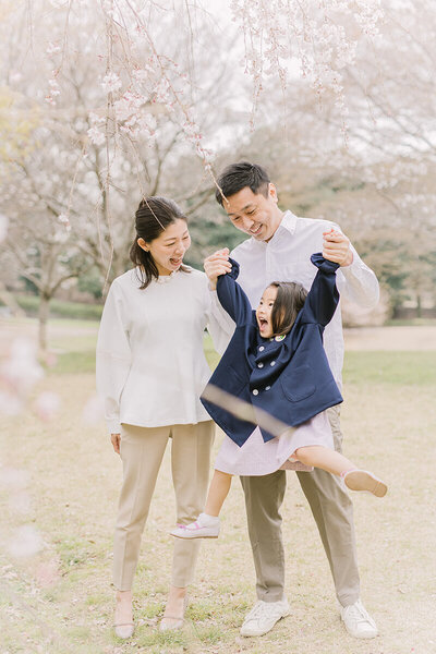 3歳女の子の入園記念の撮影で、桜の木の横でママとパパの3人で撮影。女の子はジャンプをしてパパママと目を合わせて嬉しそうにしている、とても可愛らしくオシャレな家族写真。相模原市でYukika Ishikawa Photographyが撮影。