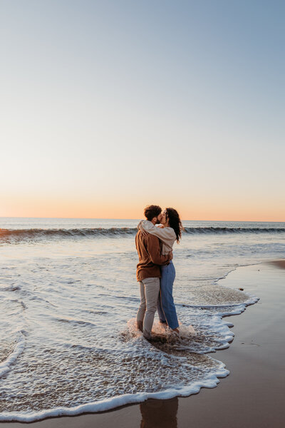 Couple during their engagement photoshoot with waves crashing around their legs as they share a kiss. Photo by Acacia and Grace Photography