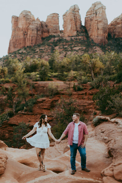 man and woman stand together with red rocks behind them for engagement photos
