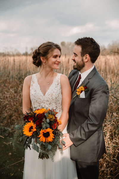 bride and groom look at each other in front of tall grasses in a fall wedding with flowers of sunflowers