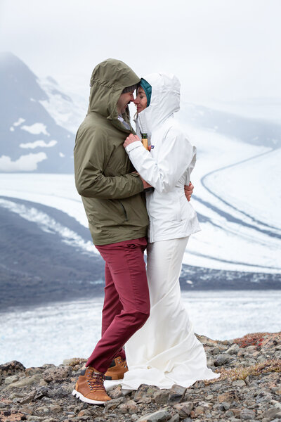 Bride & groom cozy up and embrace  high on  a mountain ridge with two glaciers merging in the background.