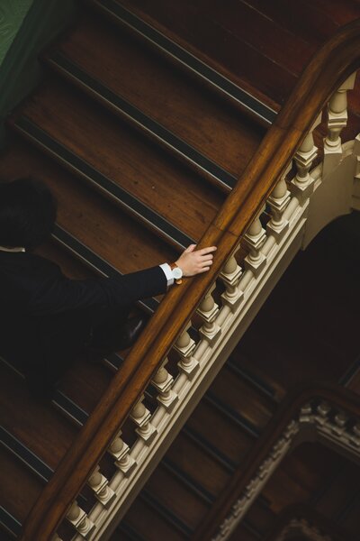 A man in a fine suit walks up a rich wooden staircase.