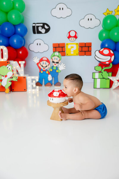 Baby wearing a blue diaper cover, leaning forward to eat a mushroom-shaped cake.
