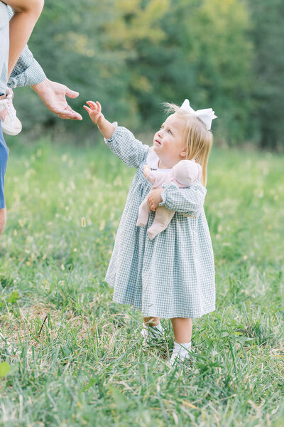Toddler girl holds baby doll and reaches for dad's hands