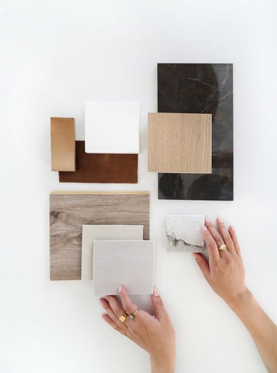 white desk top, material samples: wood, black tile, white and creamy tiles, brown leather, two hands with gold rings touching tiles