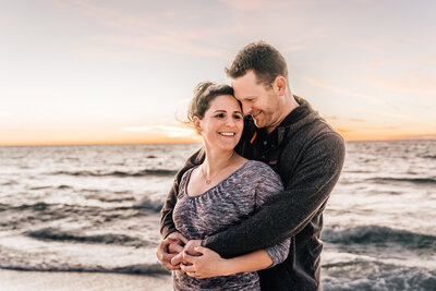 Kate and Nick hug one another with a sunset and waves behind them.