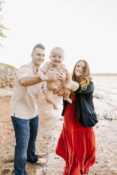 Family of three swinging baby boy back and forth like an airplane during their family session at the lake in aiken sc