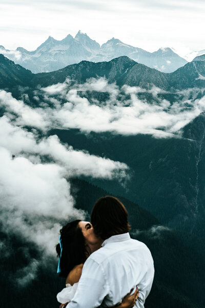 a couple in wedding attire stand in front of jagged, blue mountains as white, puffy clouds roll in. The groom gazes out as his bride kisses his cheek. Taken from a higher vantage point behind them by their adventure elopement photographers, we have t  chance to see what our couple is experiencing, as they view it.