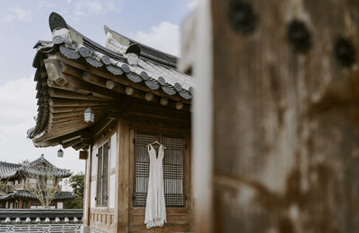 a white, beaded wedding dress hangs in front of a door to the hanok in south korea