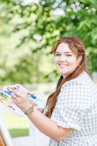 Confident live wedding artist Elyse glancing at the camera while working on a painting, embodying her approachable and professional persona