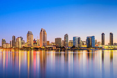 The skyline of downtown San Diego at Dusk with the lights of the building reflecting on the water with a clear blue sky background and all of the buildings starting to light up