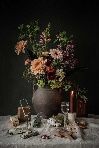 Stilleven met zalmkleurig bloemstuk in kruik van aardewerk met rondom vintage auto's, handzaag, mos, kaas en glas met jenever