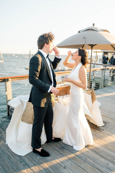 Newport RI Wedding Photographer | A couple in formal attire enjoys oysters on a sunlit pier. The woman in a white dress and man in a suit.
