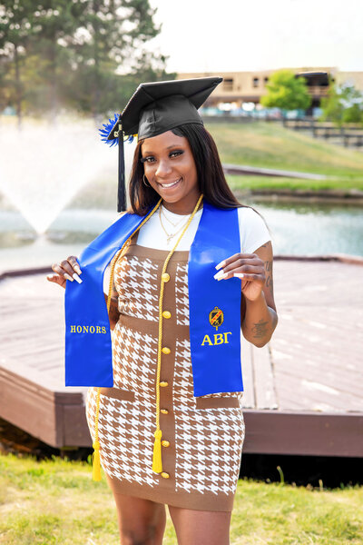 College graduate wearing black graduation cap, blue honors sash, yellow cord and brown and white dress posing in front of a pond