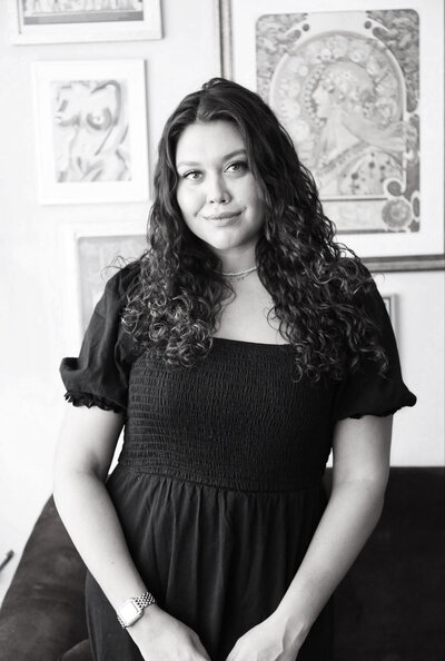 Black and white photo of woman with long, dark, curly hair wearing a black ruffled top standing in front of a gallery wall of art and softly smiling,