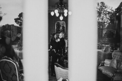 Black and white photo of a wedding photographer and videographer duo smiling at each other, captured through a window reflection. Based in the Lake District, UK, and available for destination weddings worldwide.