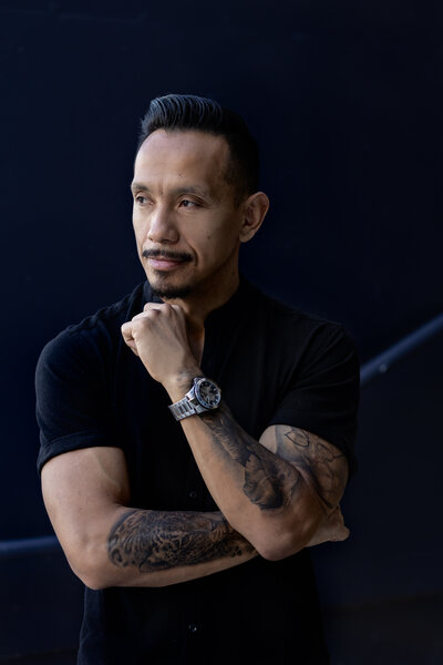 Confident man with tattoos wearing a black shirt and a stylish watch, posing thoughtfully with his hand on his chin against a dark background.