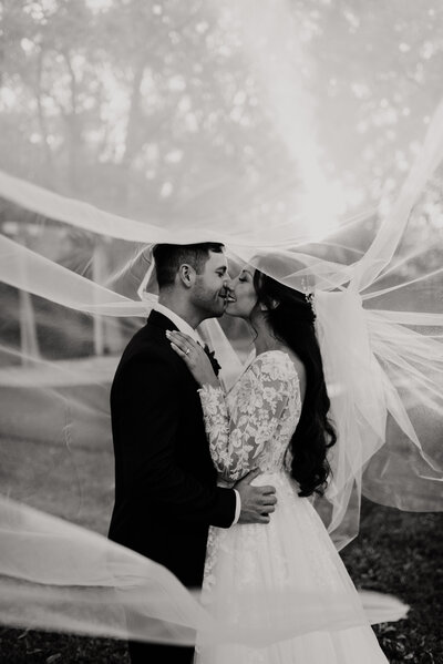Bride and groom both under long veil on their wedding day as the wind blows and they touch noses and smile