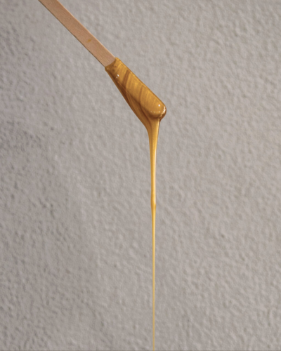 A wooden stick drips thick, golden wax against a light gray textured background.