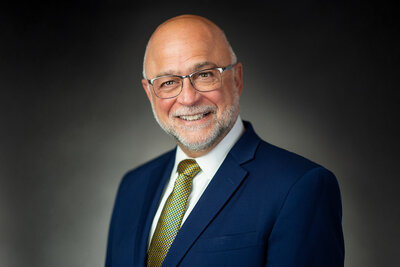 Headshot of older male executive with glasses and beard, smiling in dark suit and green tie