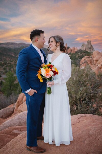 A bride and groom couple stand atop a red rock at Garden of the Gods as the sun sets behind them.