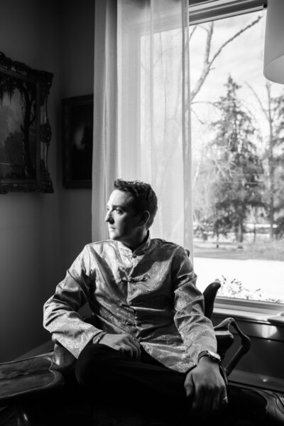 A man dressed in a formal patterned jacket sits by a window, gazing outside thoughtfully as natural light illuminates his face in a black and white portrait.