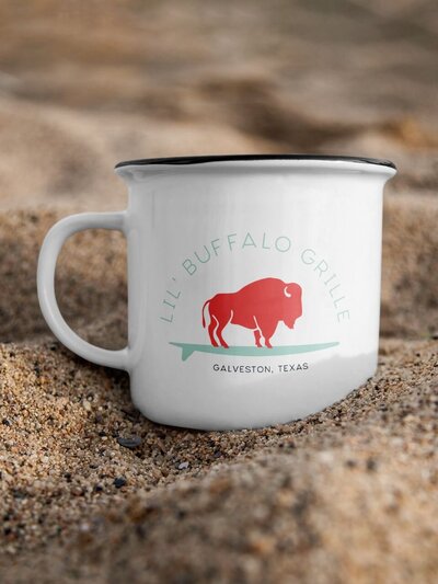 White enamel mug with a coral buffalo and mint surfboard logo for Lil' Buffalo Grille, placed in sandy beach setting with “Galveston, Texas” printed below the mark.