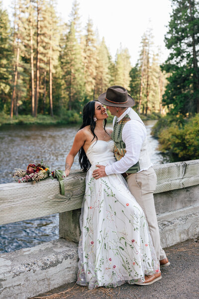 A native American bride wearing a strapless white gown with floral applique gazes into her groom's eyes as she leans back onto a bridge over the Metolius River. | Bend Elopement Photographer