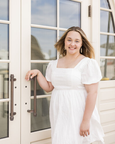high school female senior with medium length sandy blonde hair, wearing a white short sleeve dress, standing next to a set of glass doors, smiling at the camera