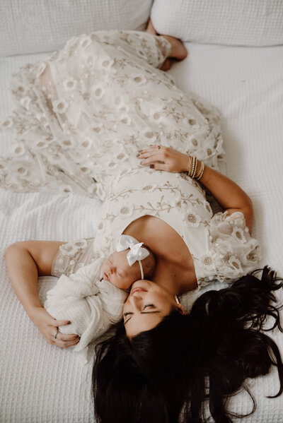 New mom in white dress laying in bed with newborn baby girl wrapped in blanket next to mom.