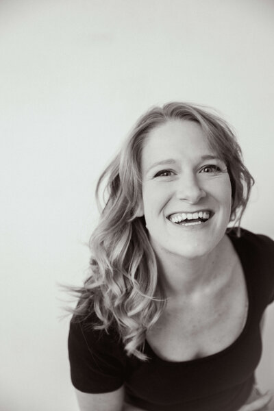 Black and white portrait of a woman with wavy hair laughing joyfully, leaning slightly forward in a candid moment against a light background.