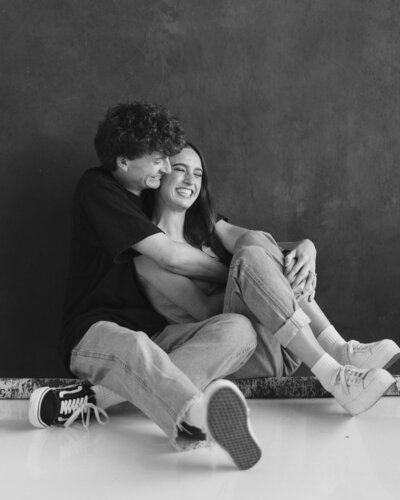 Couple sitting on the floor with their arms around each other and smiling in front of a dark studio background