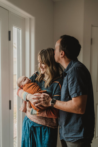 mom holding newborn baby, being held by her husband who is kissing her next to their porch door