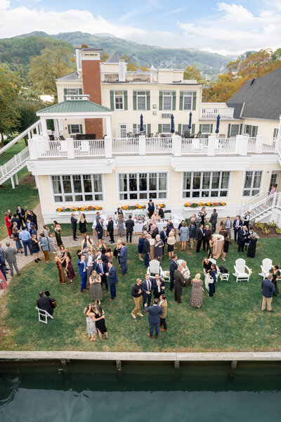 Aerial view of a formal outdoor gathering by a waterfront. Guests in suits and dresses mingle on a lawn with white chairs and tables. A large building with balconies overlooks the event. Hills and trees are visible in the background.