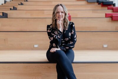 Woman sitting on wooden steps, smiling, wearing black floral blouse and black pants.