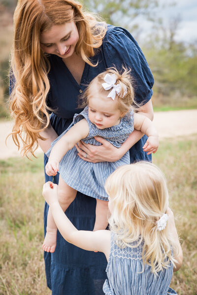 a mom holding a baby and sisters looking at a flower