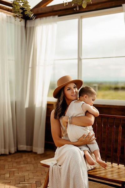 Mom holds toddler son while sitting on a bench in a greenhouse