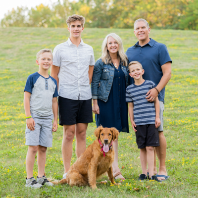Family portrait outdoors with two adults, three boys, and a golden retriever dog, standing together in a grassy field.